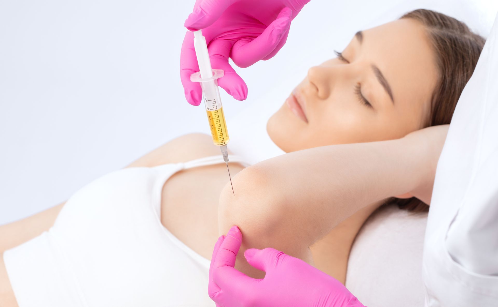 Gloved clinician injecting a patient's upper arm while she lies relaxed on a white treatment bed