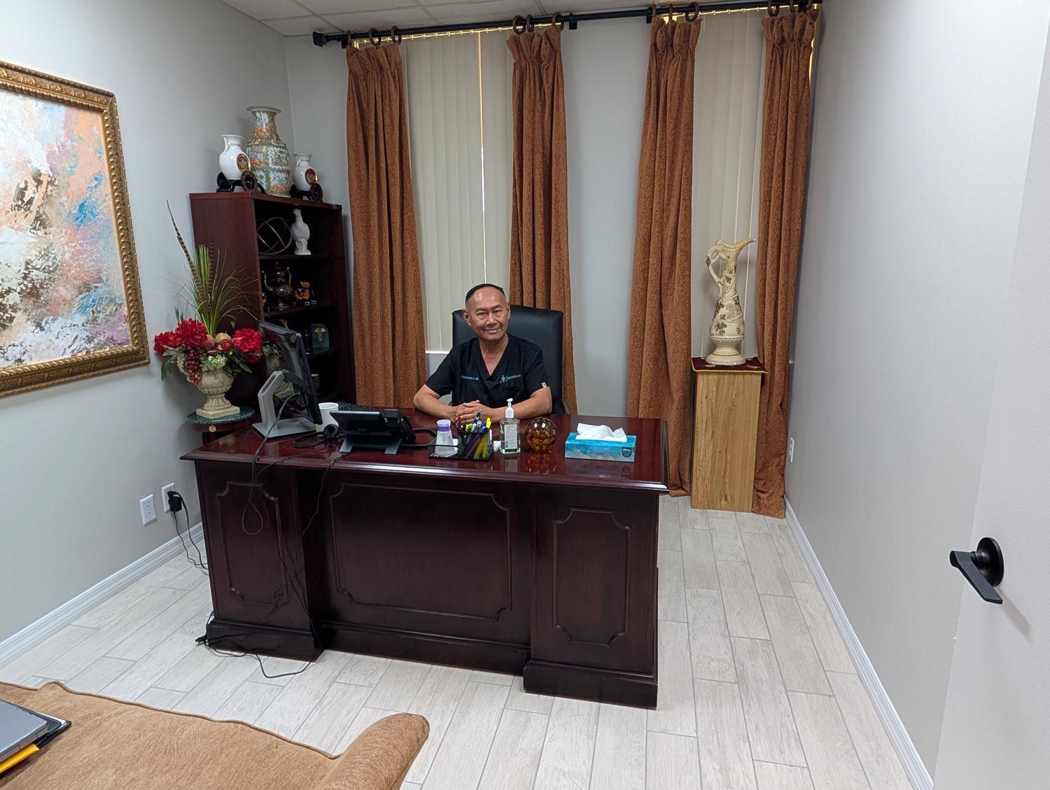 Man seated behind a dark wooden desk in a small office with curtains, wall art, and a vase of flowers.