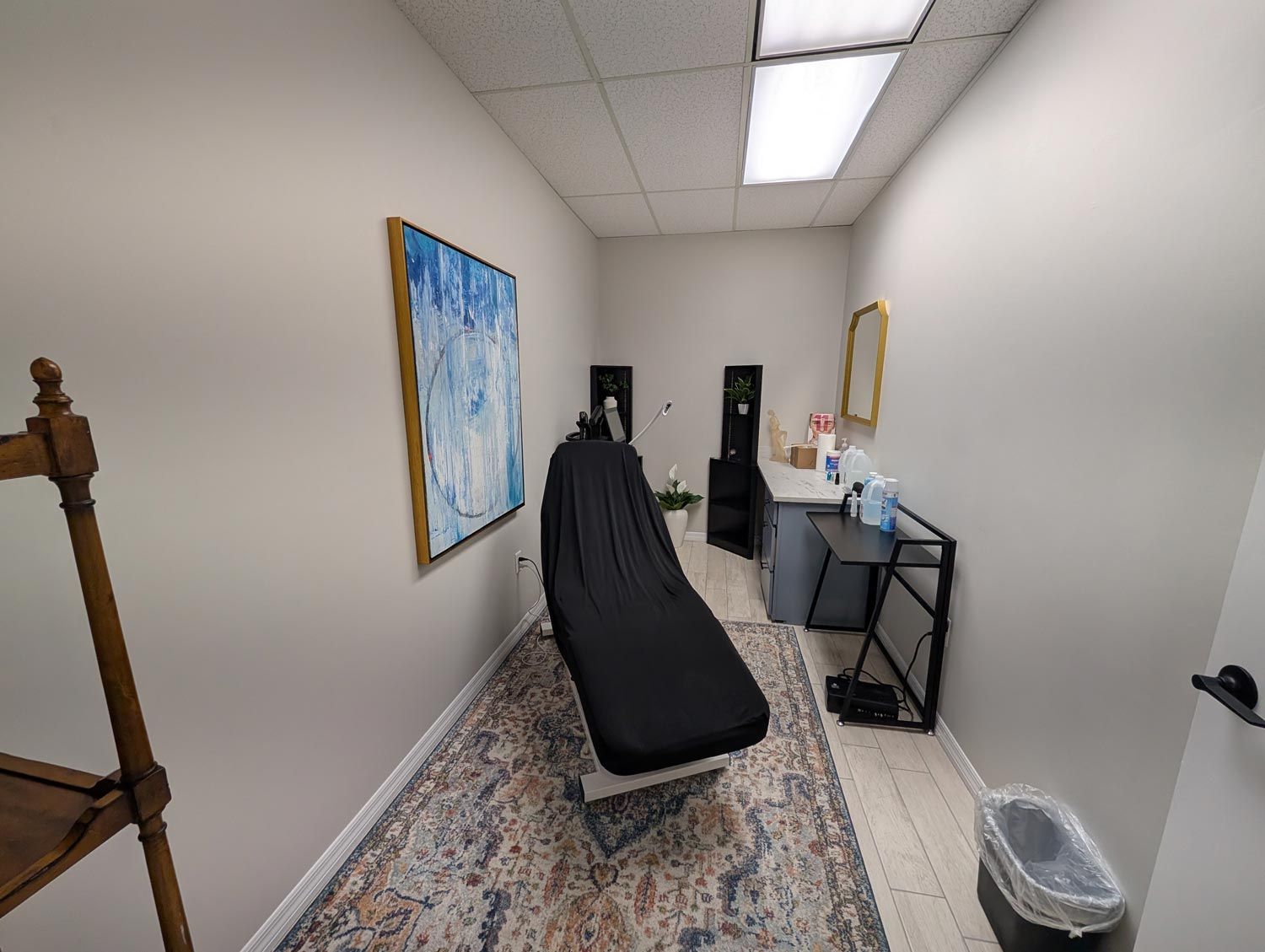 Small office room with a black massage table, colorful wall art, desk, and trash can on a patterned rug.