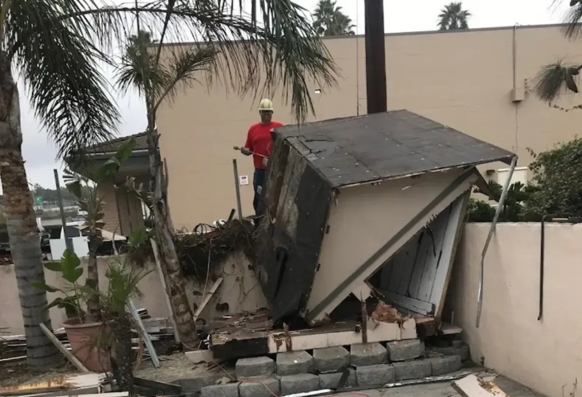 A man in a hard hat stands next to a shed that has fallen over