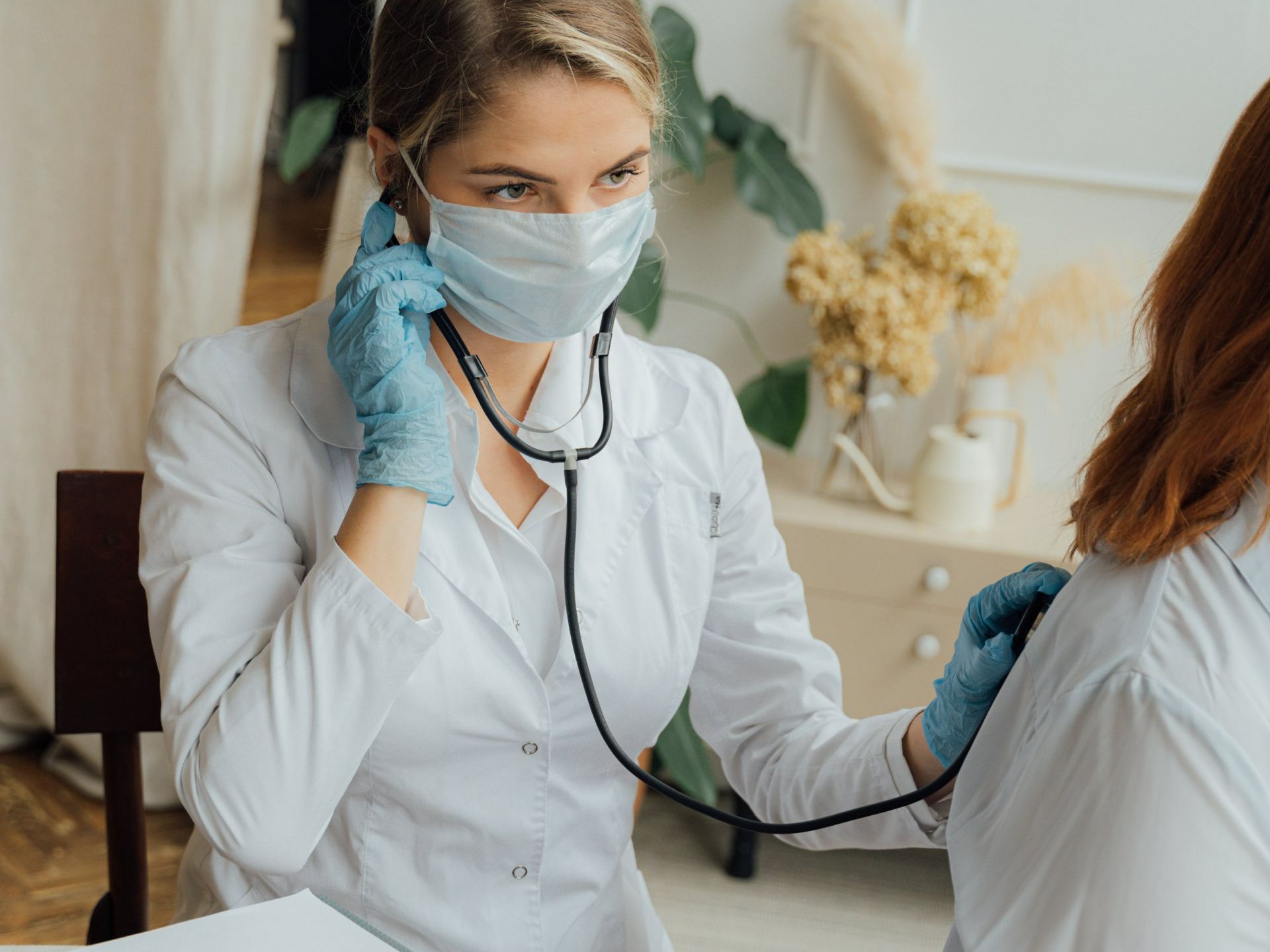 A doctor wearing a mask and gloves is listening to a patient 's heartbeat with a stethoscope.