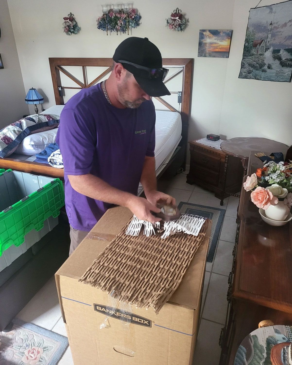 Man in purple shirt opening a cardboard box on a bed in a bedroom