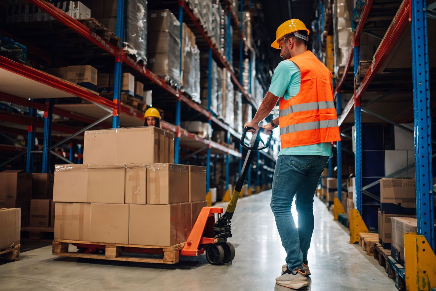 Warehouse worker in orange vest moving stacked boxes with a pallet jack in a warehouse aisle