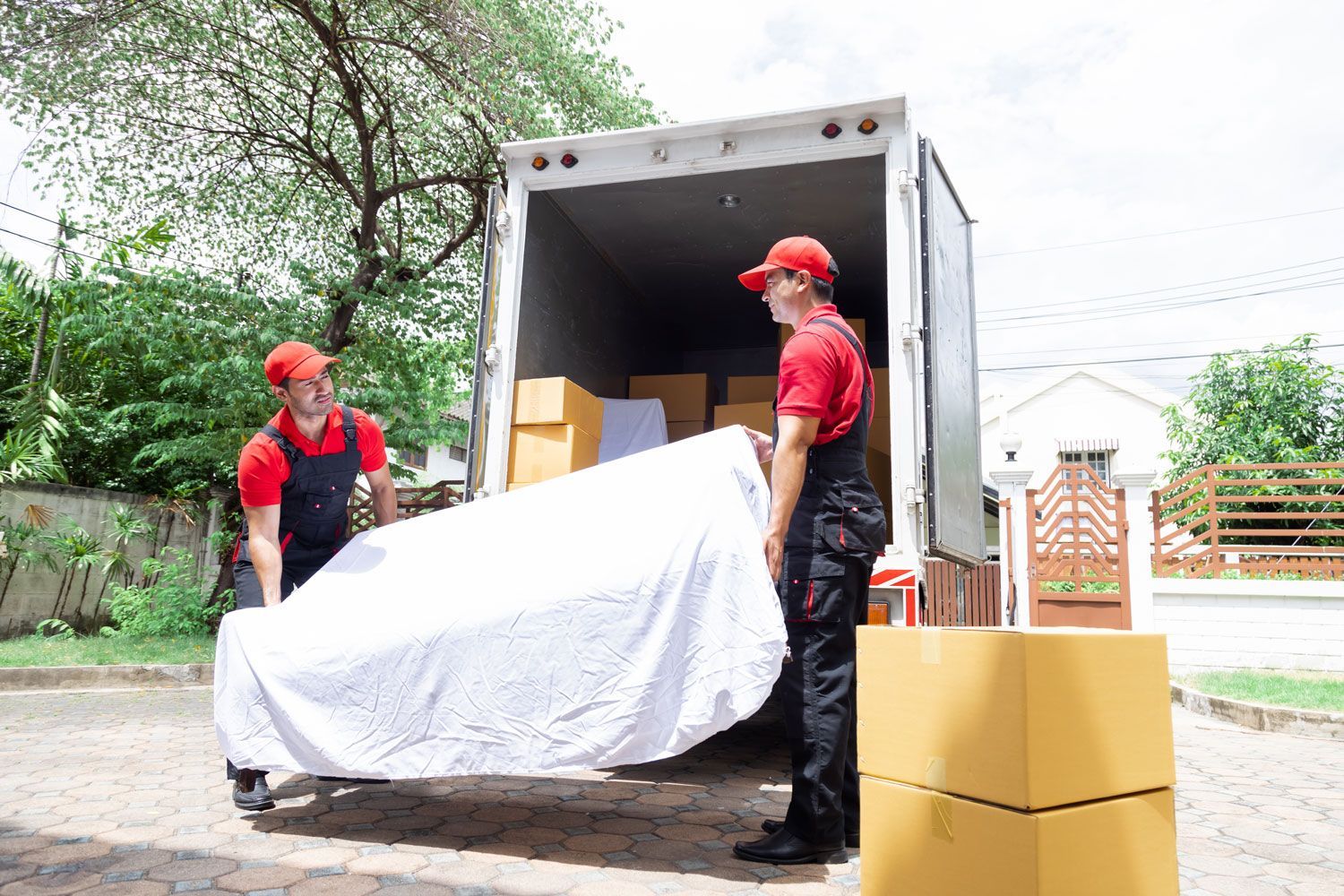 Two workers in red uniforms unload a white-covered item from a truck beside stacked cardboard boxes.