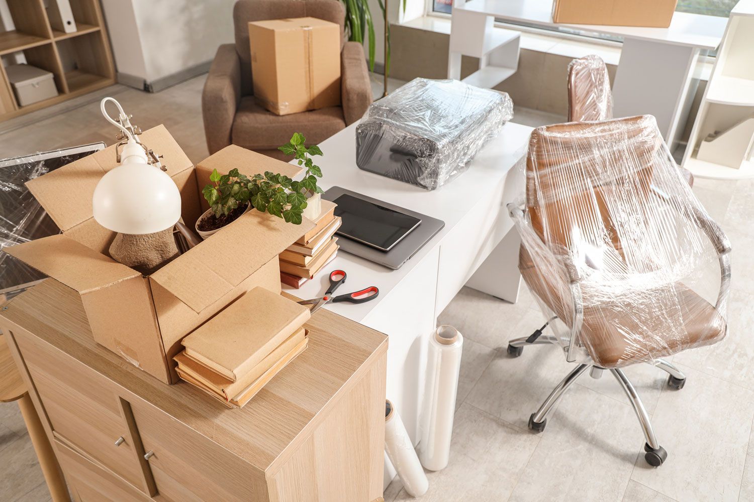 Cluttered home office with moving boxes, desk lamp, plants, and a chair wrapped in protective plastic.