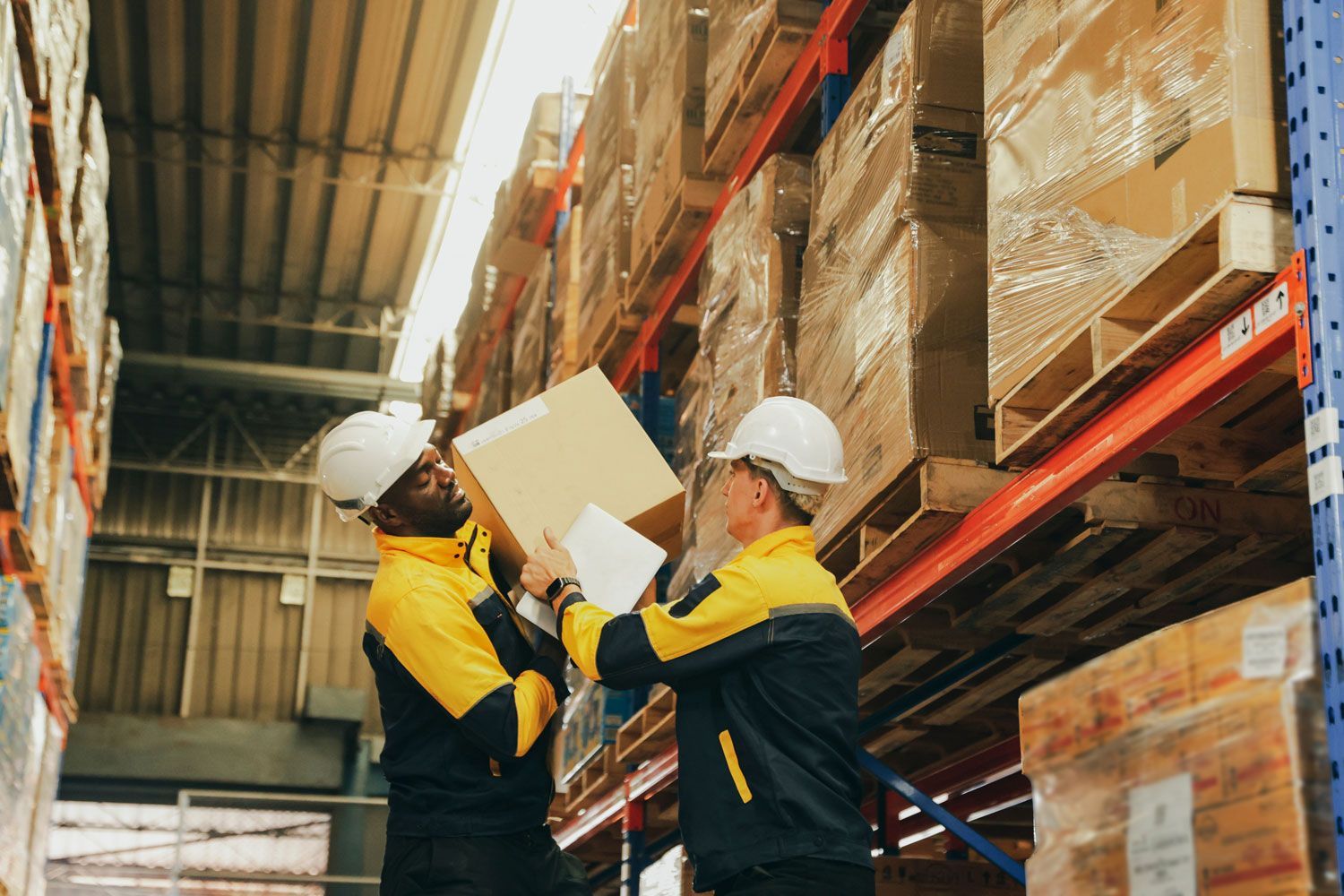 Two warehouse workers in hard hats inspecting a box on shelving aisle