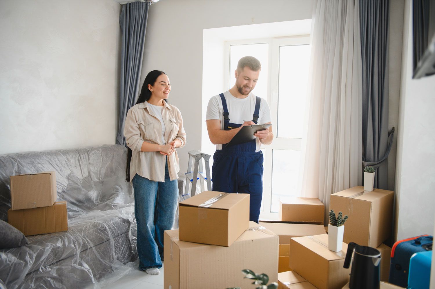 Two people unpacking moving boxes in a bright living room with a sofa and large windows.