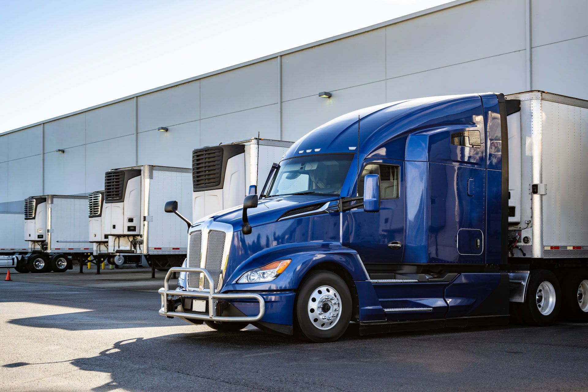 Blue semi-truck parked outside a warehouse loading dock with other trucks nearby