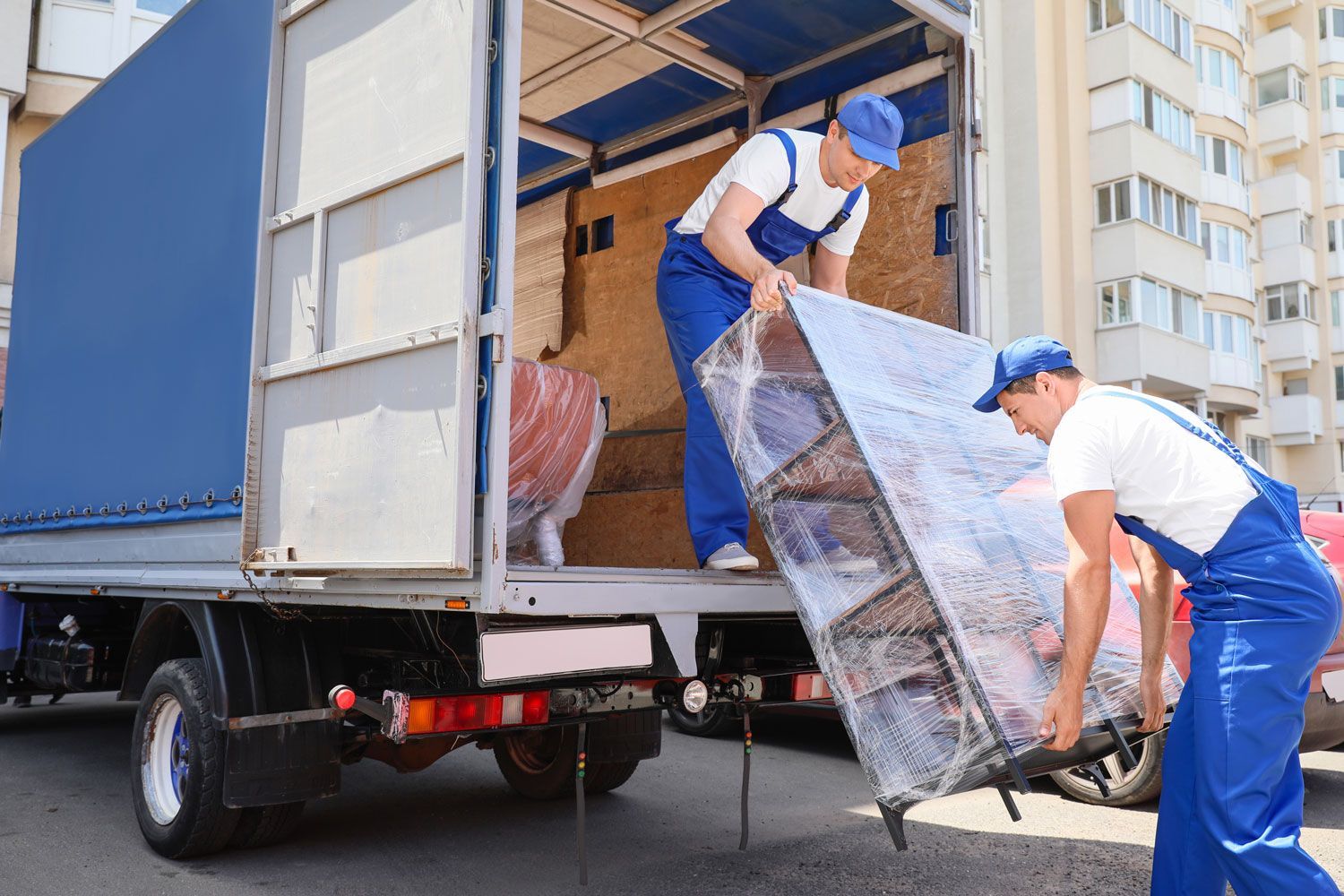 Two movers in blue uniforms unload furniture from a truck onto a city sidewalk.