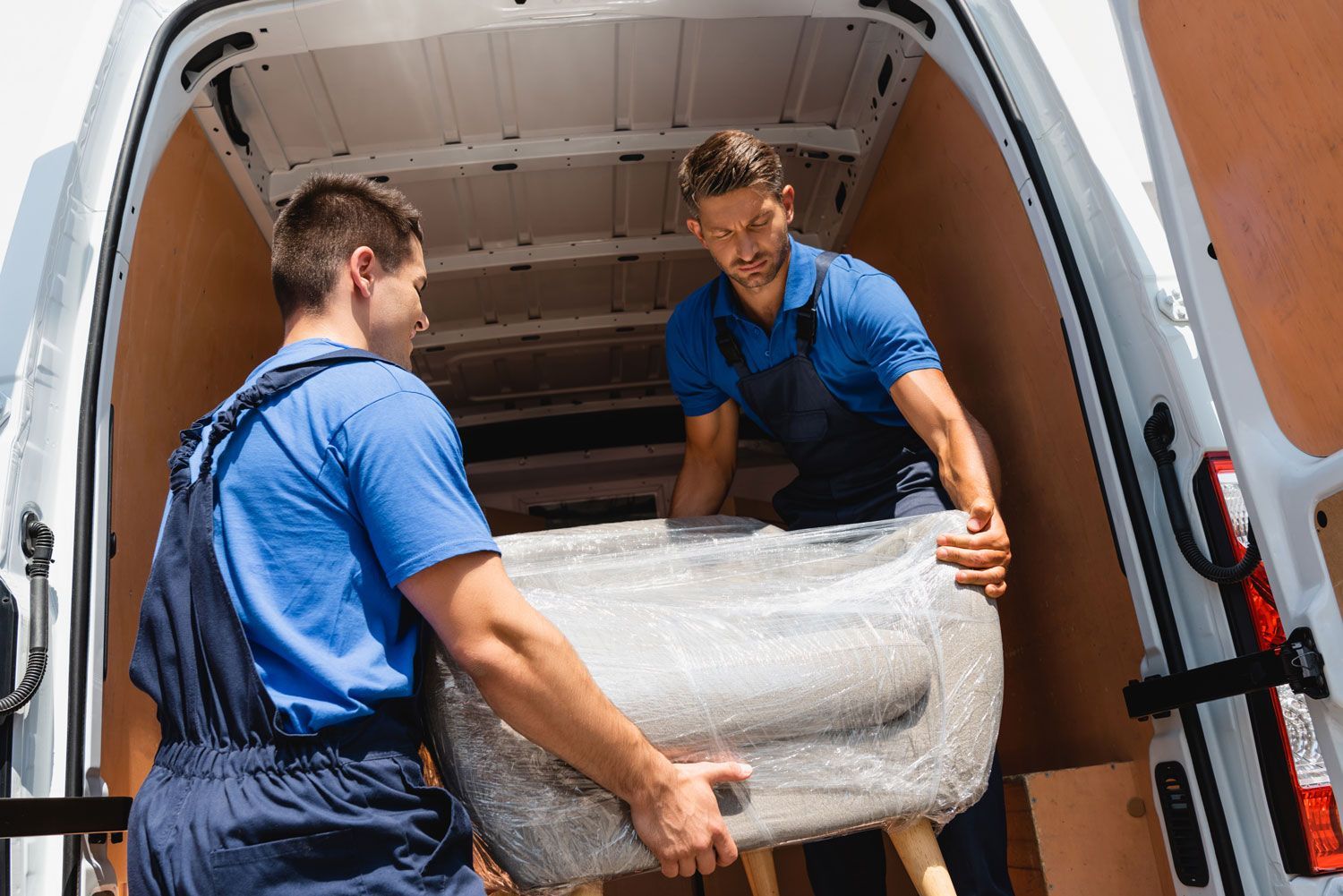 Two movers in blue uniforms carrying a wrapped appliance into a van