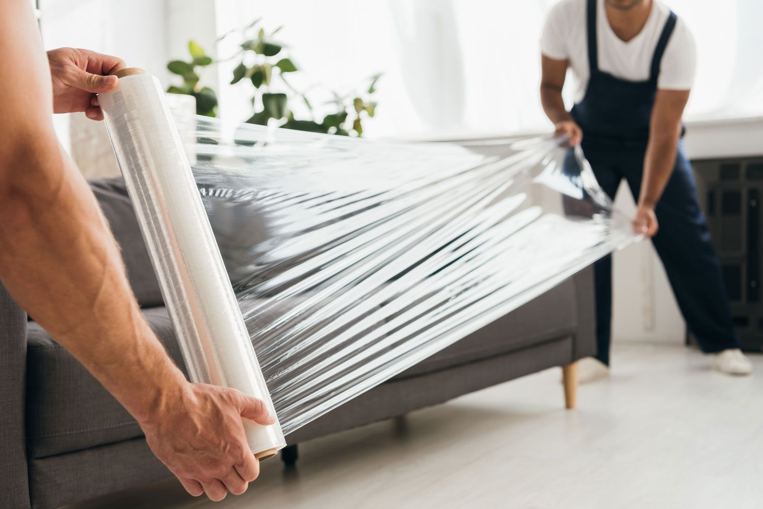 Two people wrapping a sofa in clear plastic for moving or storage