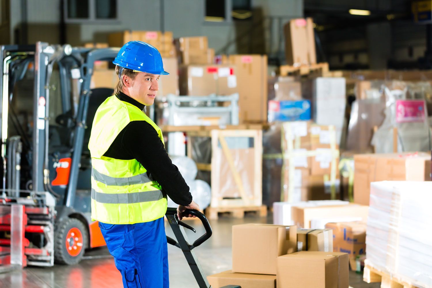 Warehouse worker in blue hard hat and reflective vest operating a pallet jack among stacked boxes