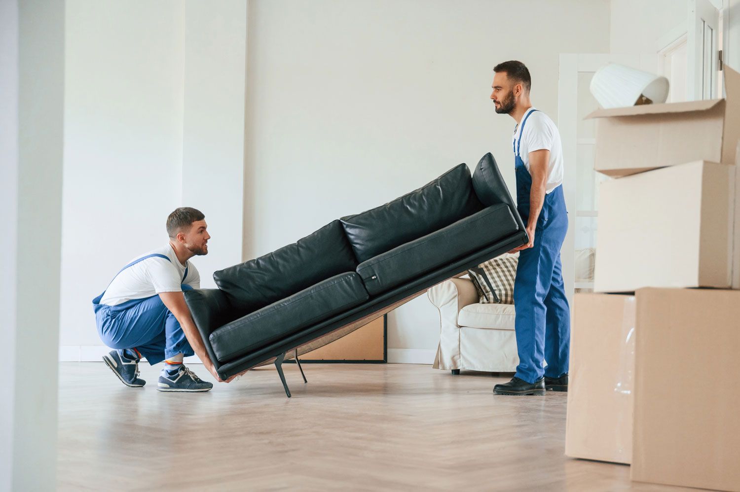 Two movers carrying a black couch through a bright, empty room with moving boxes