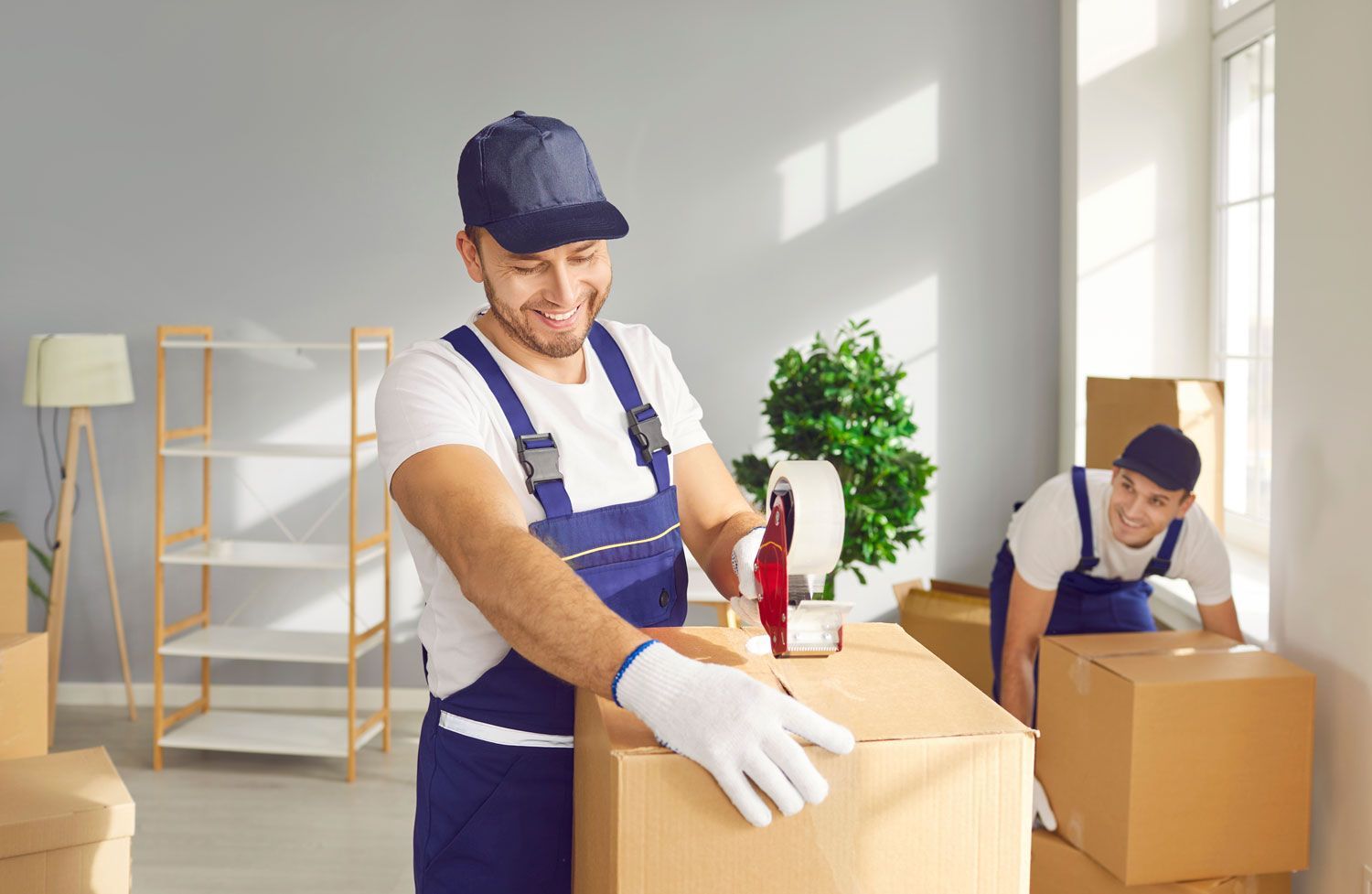 Two movers in blue uniforms packing cardboard boxes in a bright room