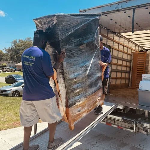 Two movers carrying a wrapped appliance into a truck bed on a sunny street