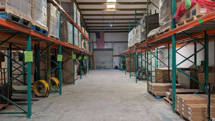 A warehouse aisle with tall metal shelving, stacked boxes, and pallets under a high ceiling.