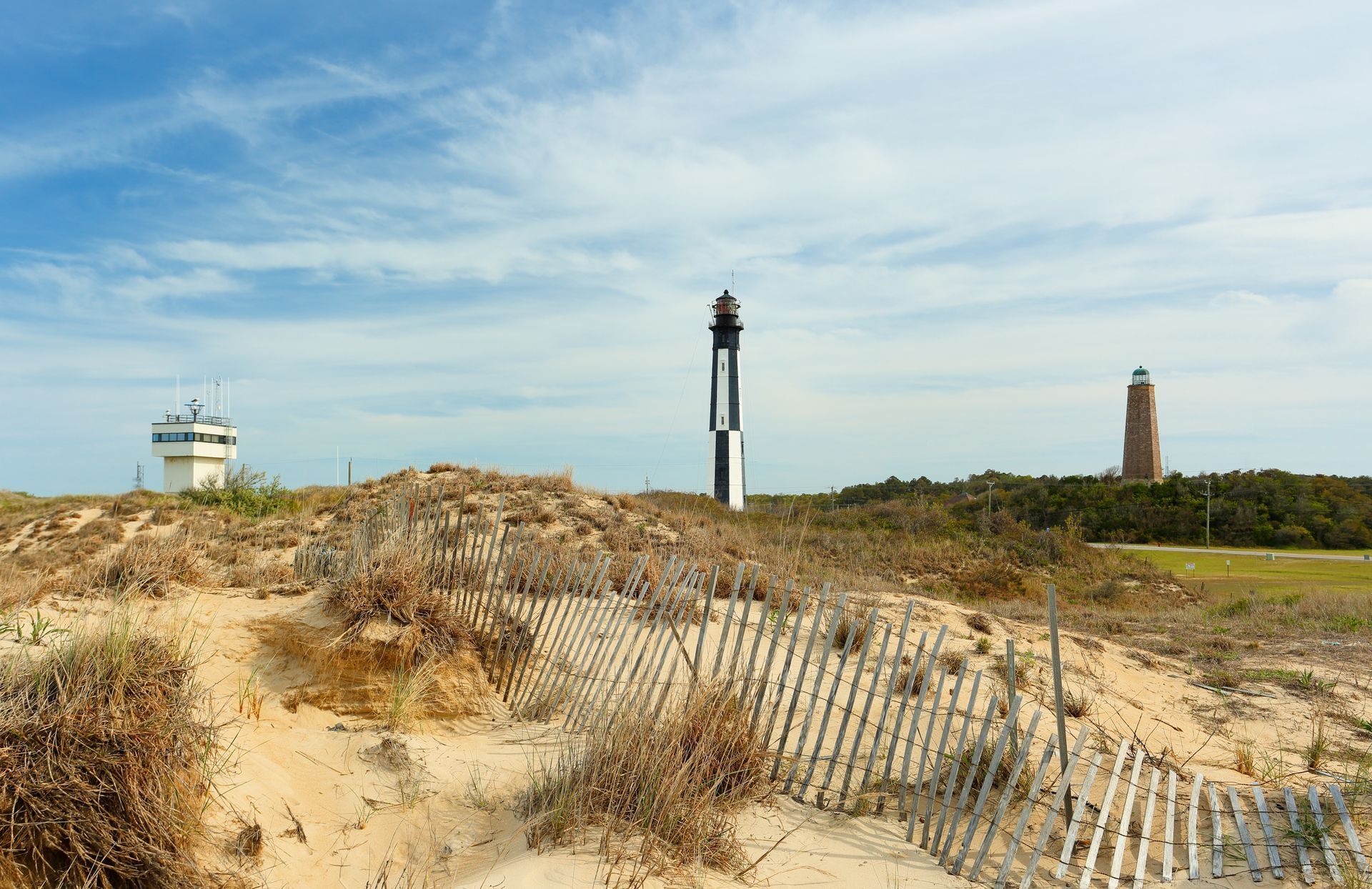 A lighthouse is sitting on top of a sand dune next to a fence.