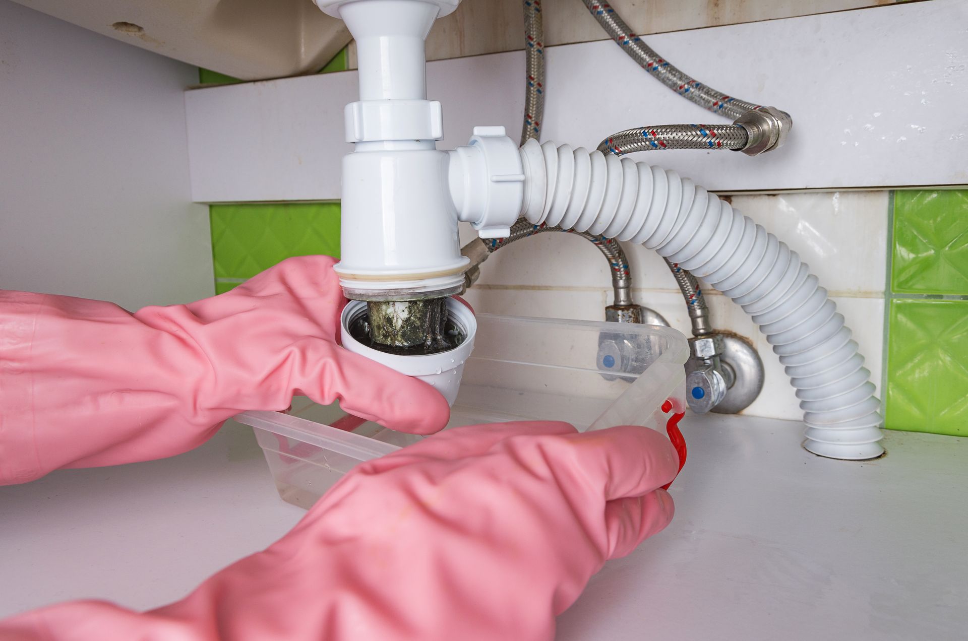 Pink-gloved hands working under a kitchen sink, adjusting the drain pipe and hose connections