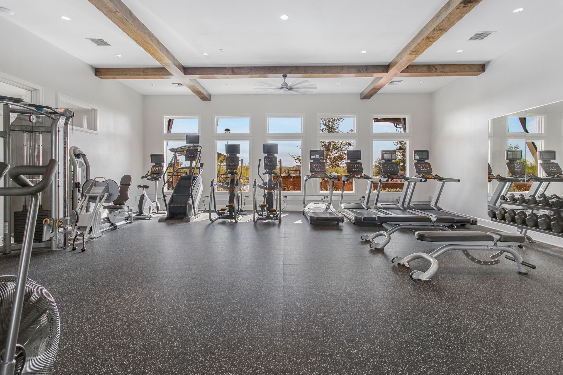 Spacious gym with various exercise machines, large windows, and a dark speckled floor. Wooden beams adorn the ceiling.