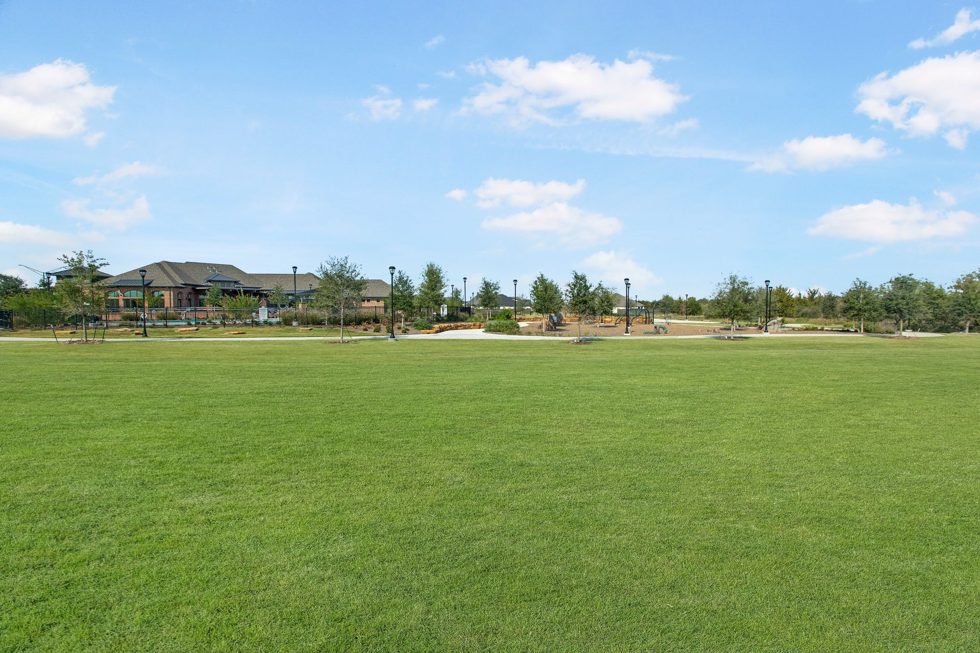 Lush green lawn with a park and buildings in the background under a blue sky with sparse clouds.