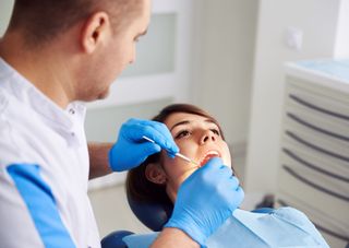 Dentist examining a woman's teeth in a dental chair; both are in a bright office, dentist wears gloves.