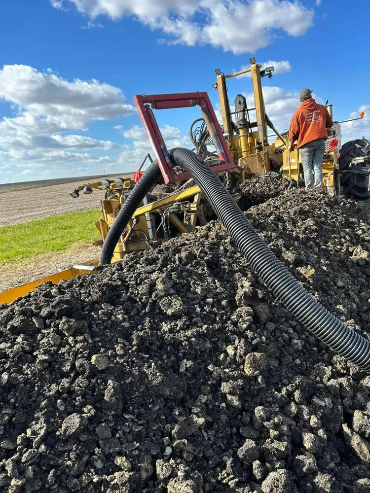 Roadwork crew and machinery on a gravel mound beside a rural road under a blue sky.