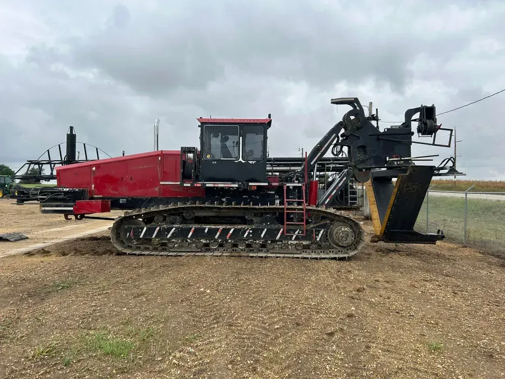 Red tracked industrial machine beside a matching trailer on a dirt field under cloudy sky