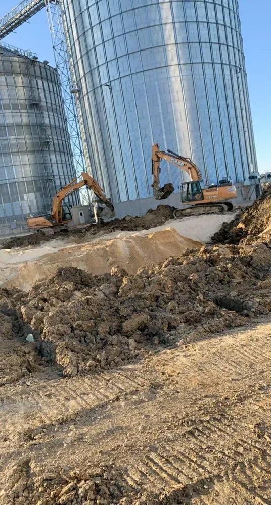 Construction site with excavators and dirt piles in front of tall silver silos
