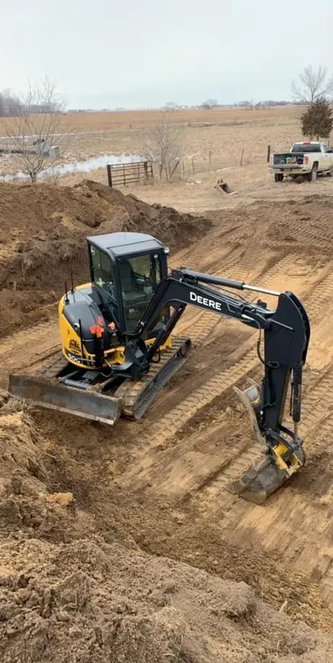 Excavator digging a dirt trench at a construction site with a dump truck in the background.