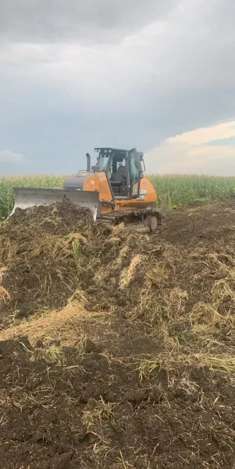 Bulldozer pushing a large pile of dirt in a crop field under a cloudy sky