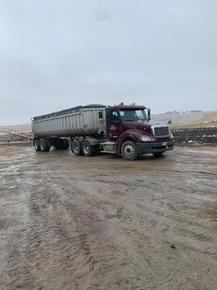 Maroon dump truck parked on a muddy construction site under an overcast sky