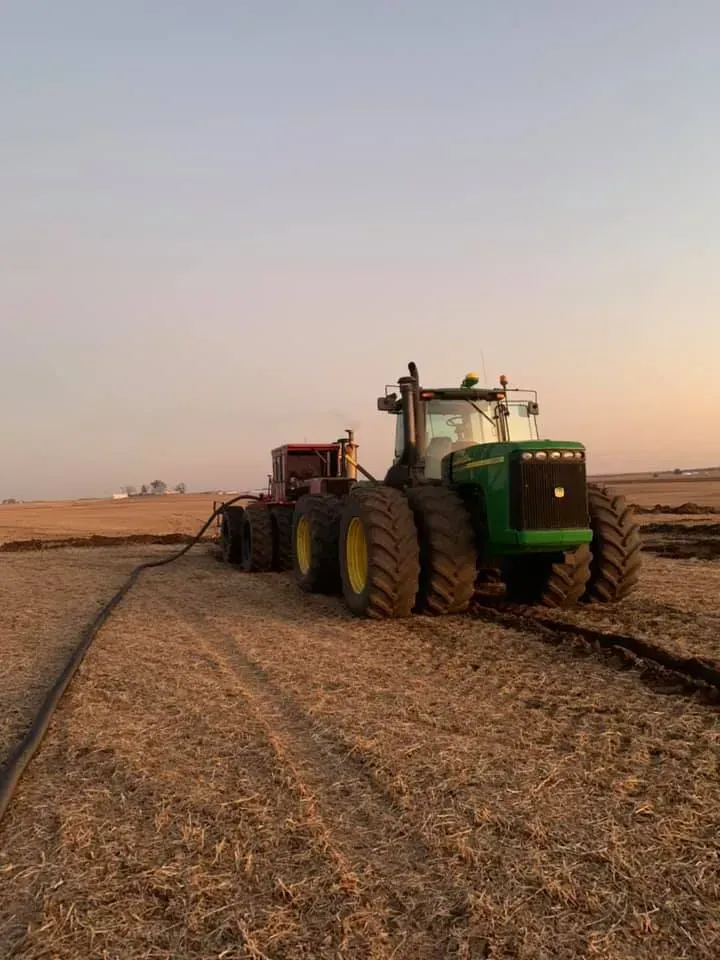 Green tractor pulling a red trailer across a dry field at sunset
