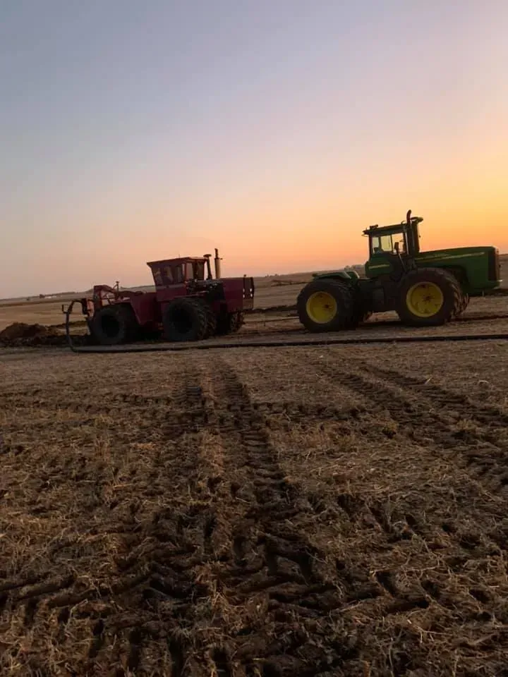 Two tractors working in a field at sunset, one red and one green.