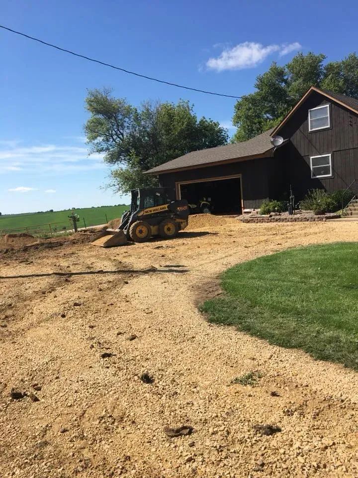 Tractor on a dirt driveway beside a barn and farmhouse under a bright blue sky