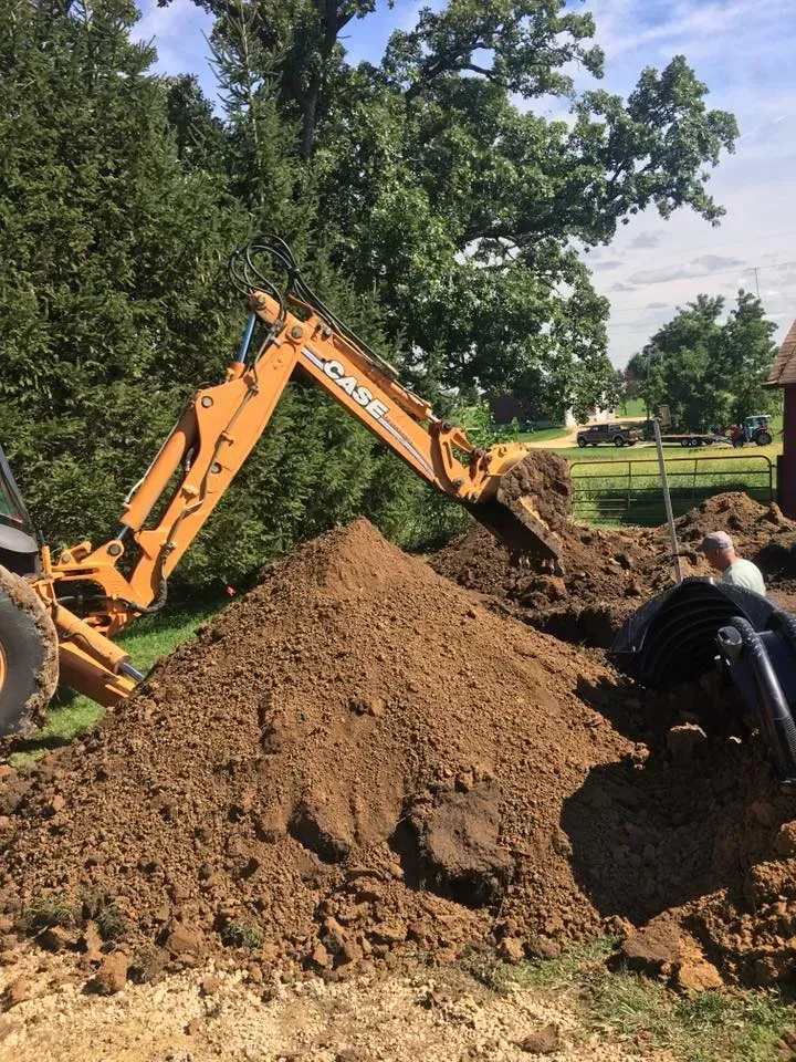 Orange excavator digging a large dirt pile in a grassy park, with a worker crouching nearby.