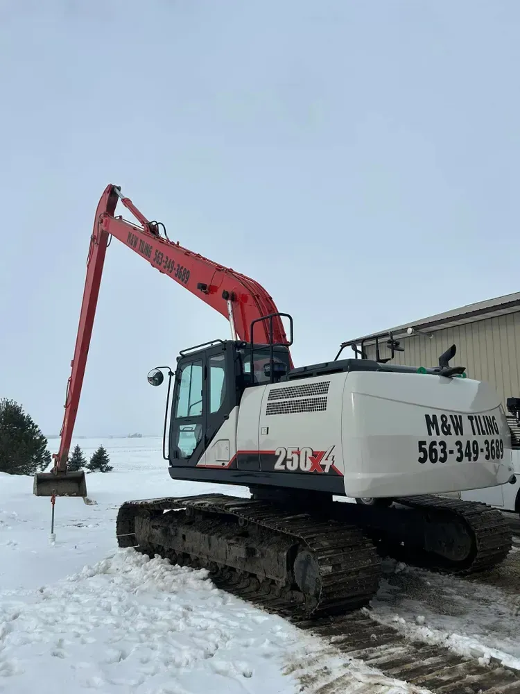 Red and white tracked excavator with extended arm in a snowy work yard beside a building