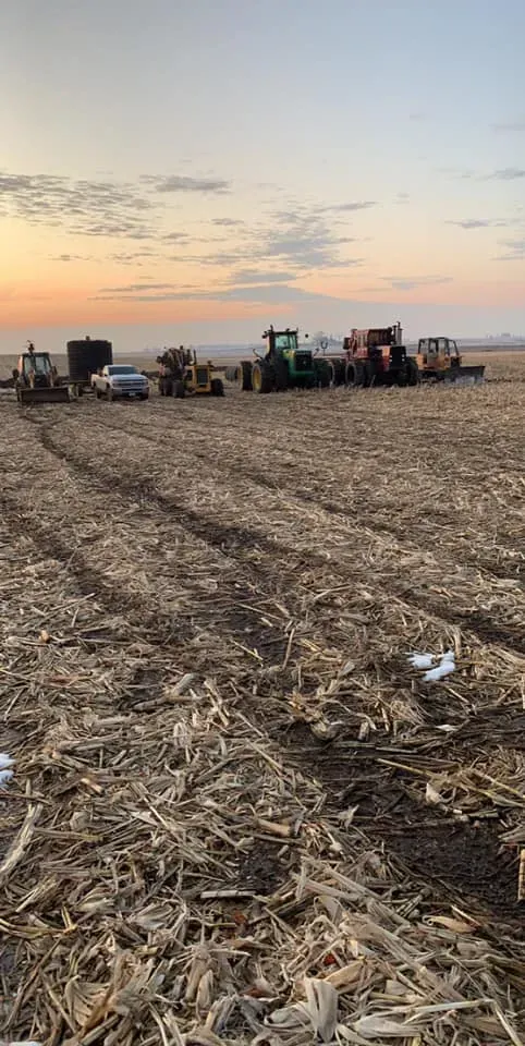 Farm field at sunset with harvested stalks and a line of tractors in the distance