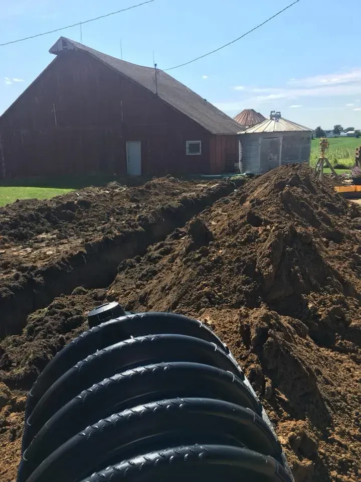 Farmyard with a red barn, large dirt mounds, and a black drainage pipe in the foreground under a blue sky