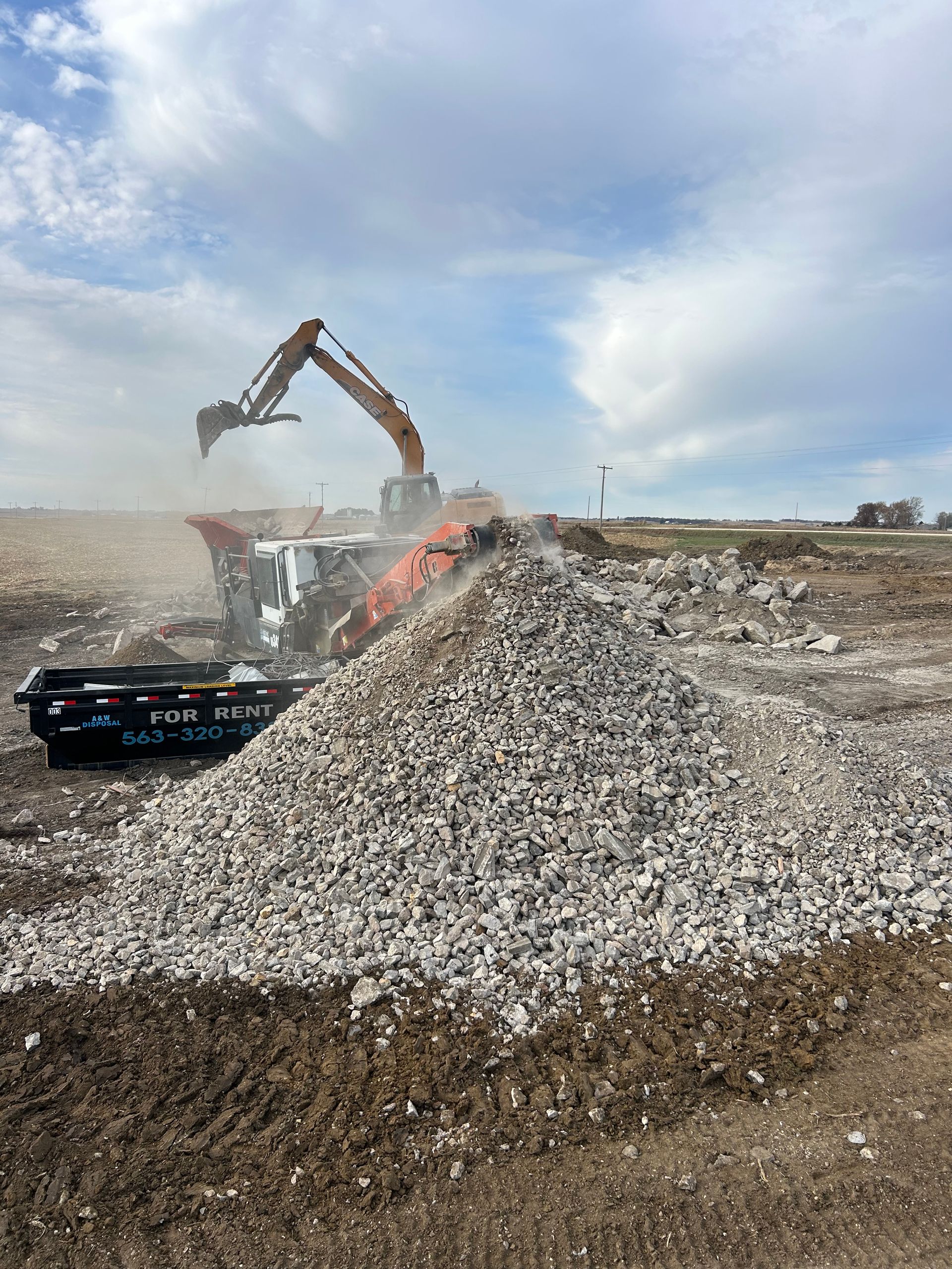 Excavator dumping rubble onto a large gravel pile at a construction site under a cloudy sky