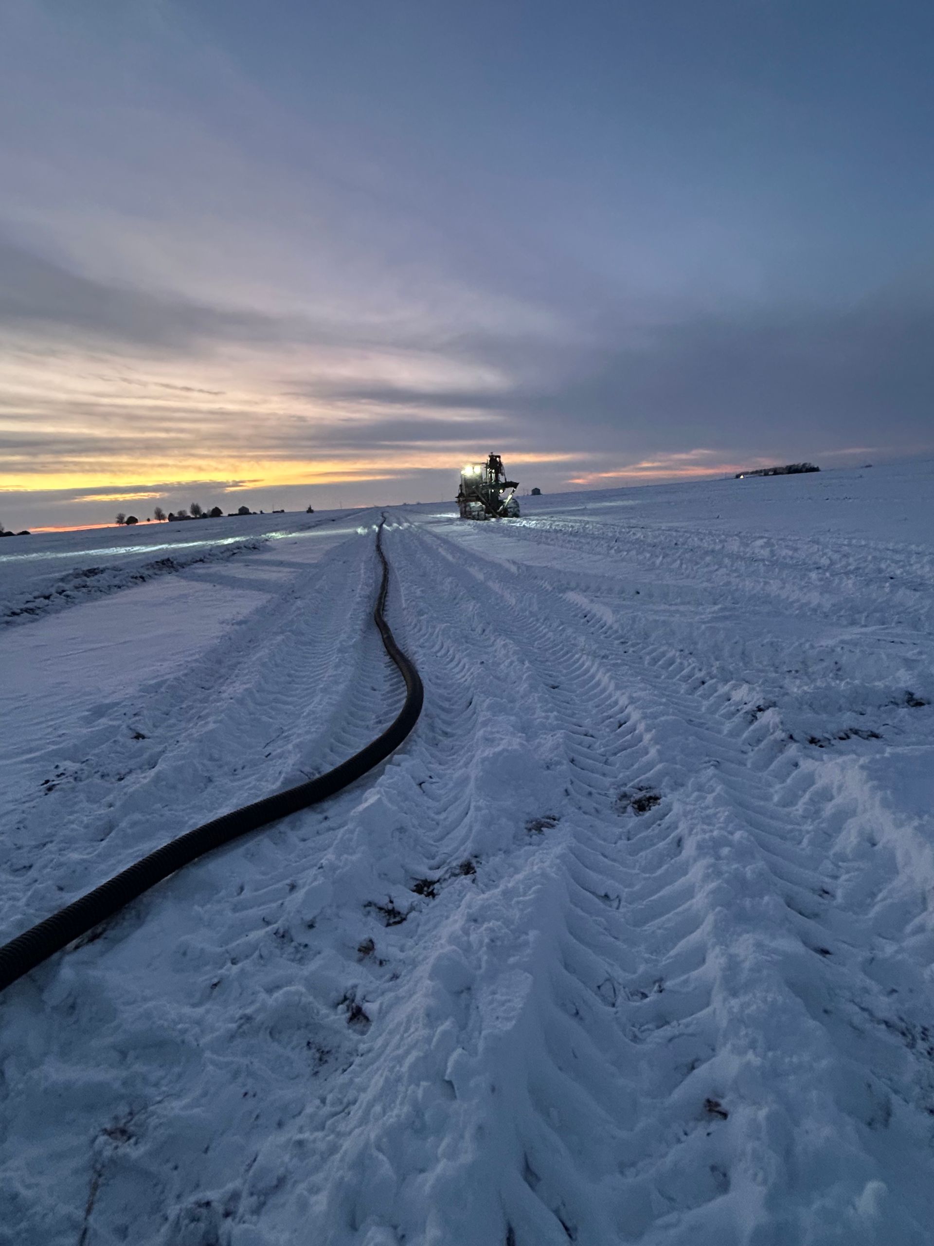 Snow-covered field at dusk with tire tracks leading to a distant tractor under a cloudy sky