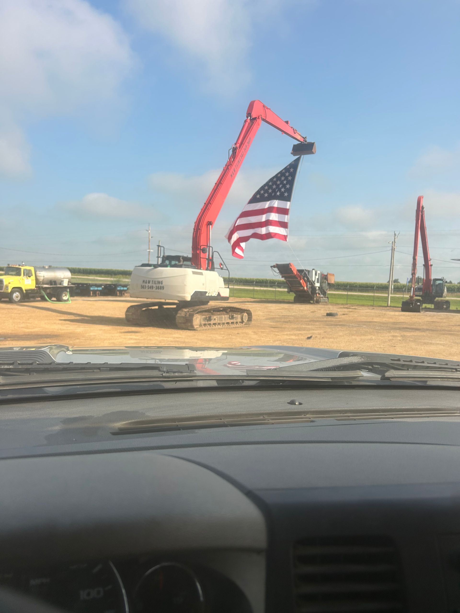 Excavator hoisting a large American flag at a construction site under a blue sky