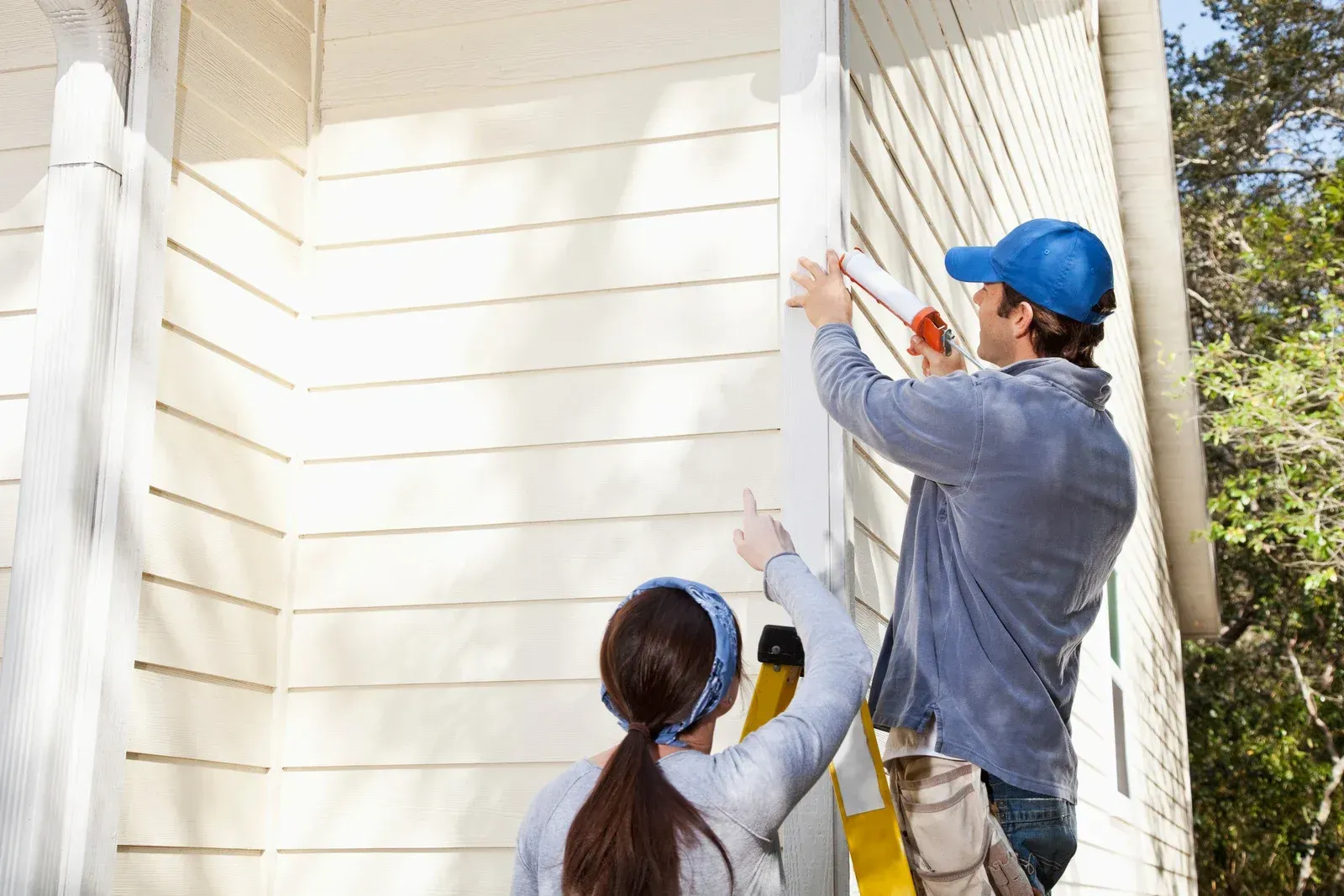 Man on ladder caulking siding as woman points; sunny outdoor setting.