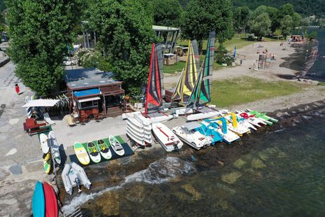 Colorful sailboats and paddleboards lined along a lake shore near trees and a sandy beach