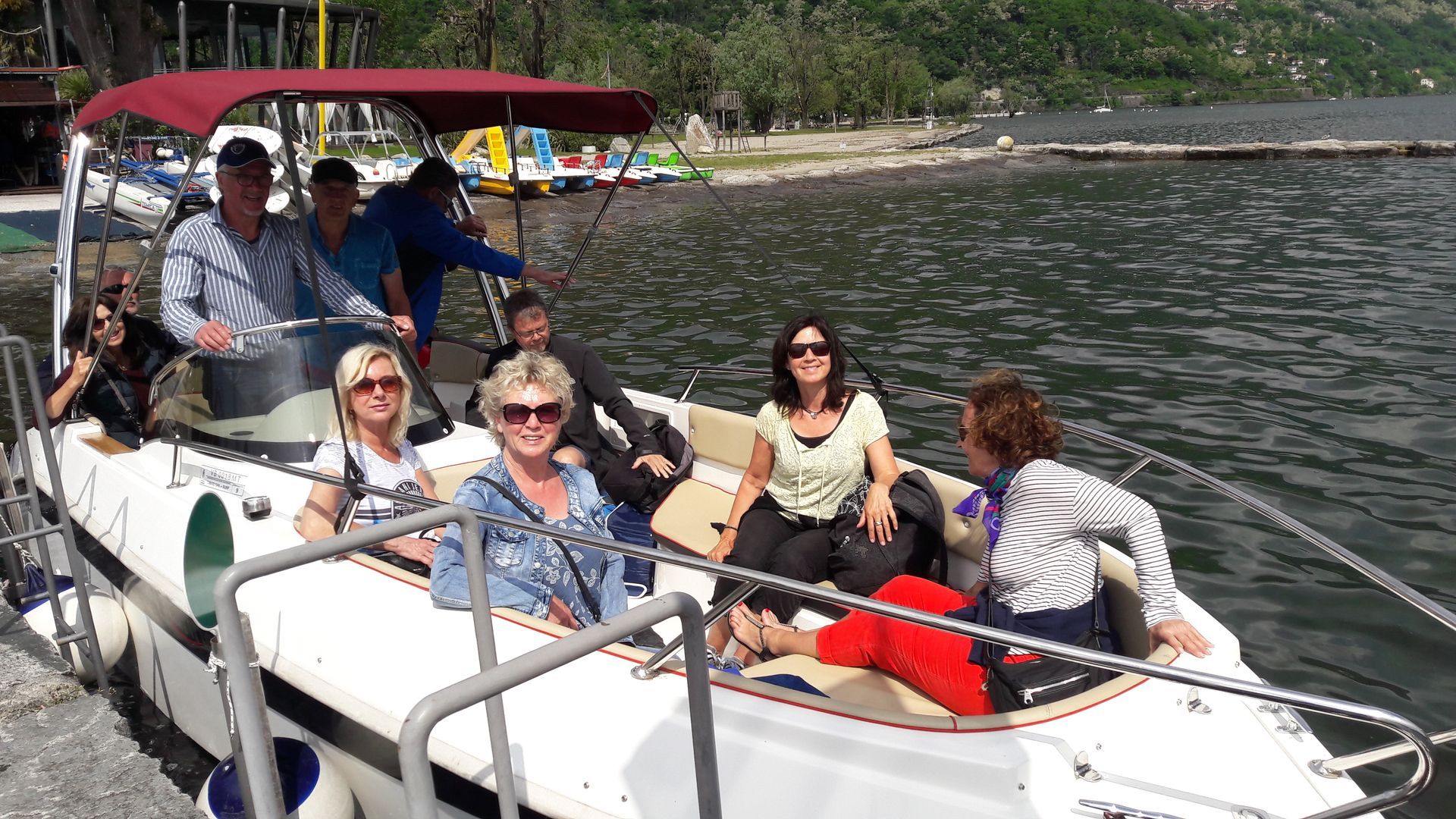 Six people on a white pontoon boat cruising on a lake, with hills and shoreline in the background.