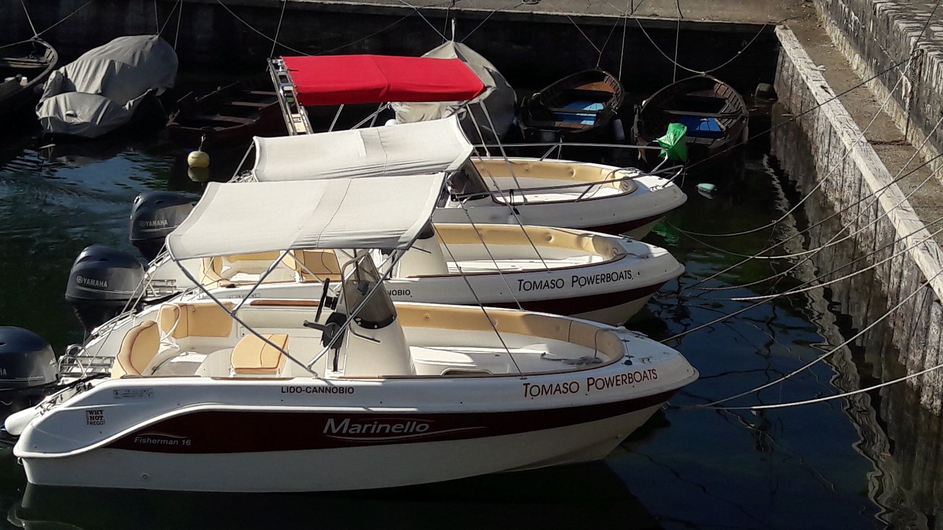 Small motorboats with red and beige canopies moored in a marina beside a stone wall.