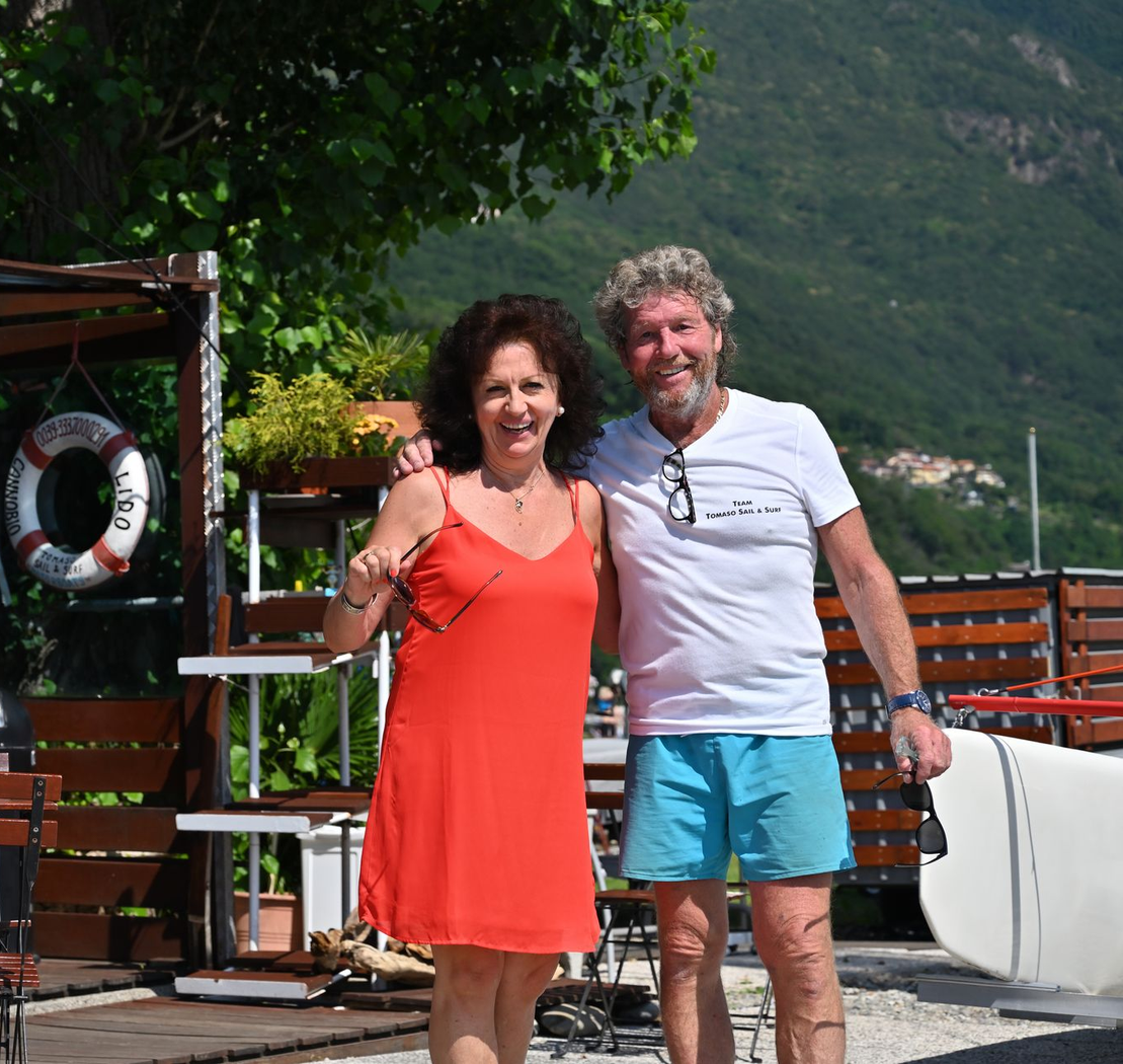 Smiling couple posing outdoors beside a waterfront, with mountains and greenery in the background