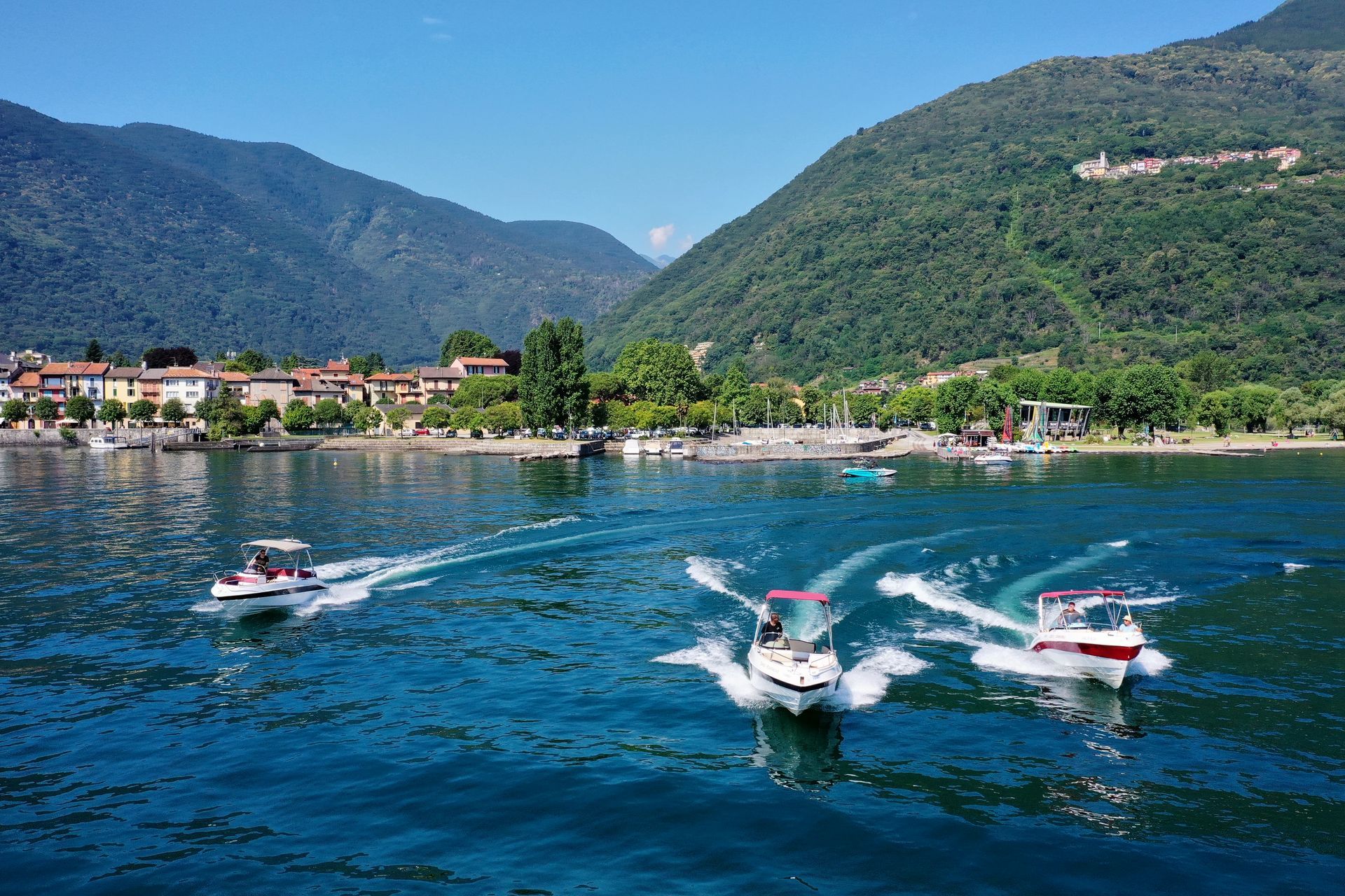 Three speedboats racing across a blue lake with mountains and a lakeside town in the background