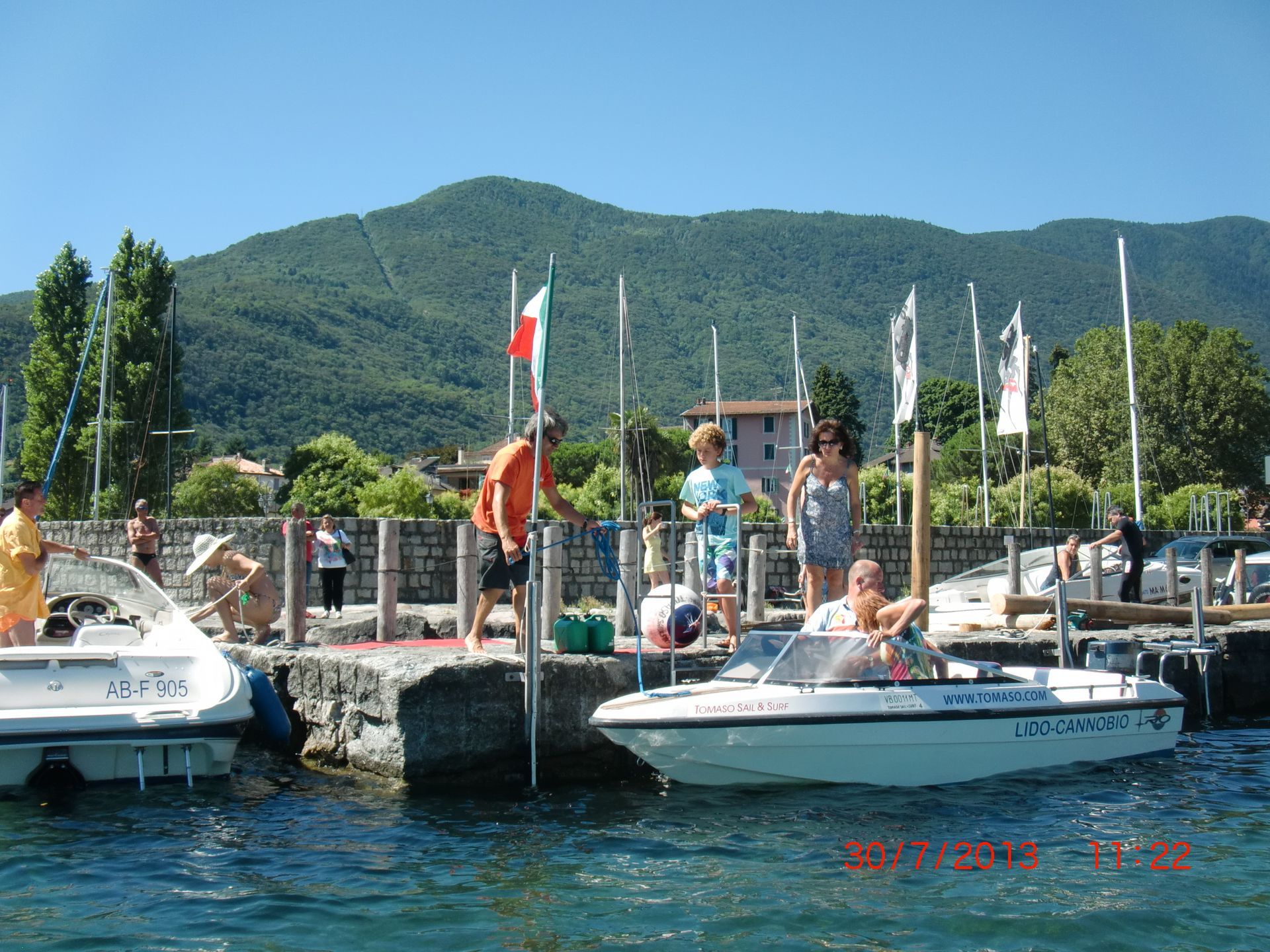 Small boat dock on a lake with people, flags, and green mountains in the background