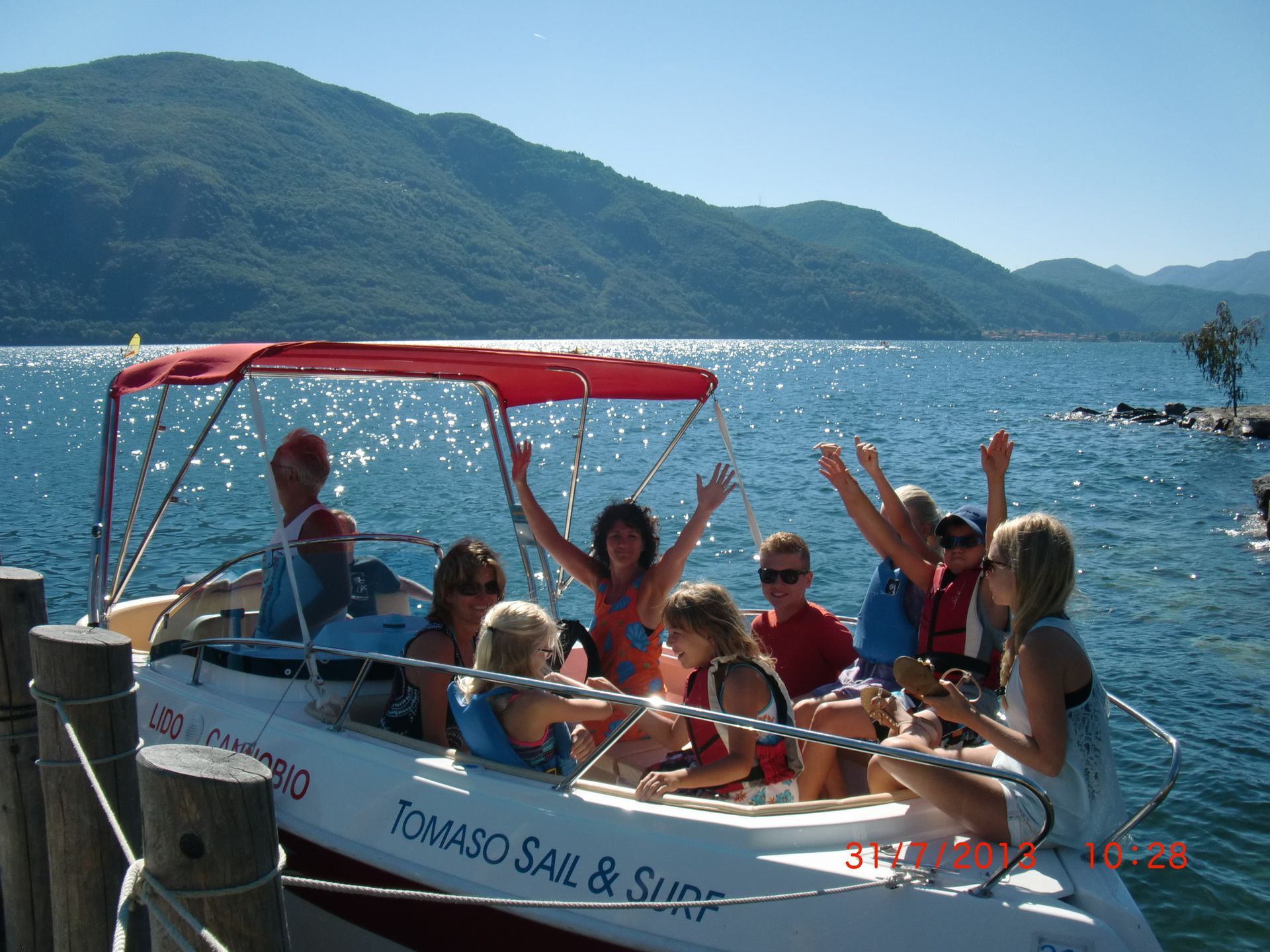 People waving on a small boat with a red canopy on a sunny lake, mountains in the background