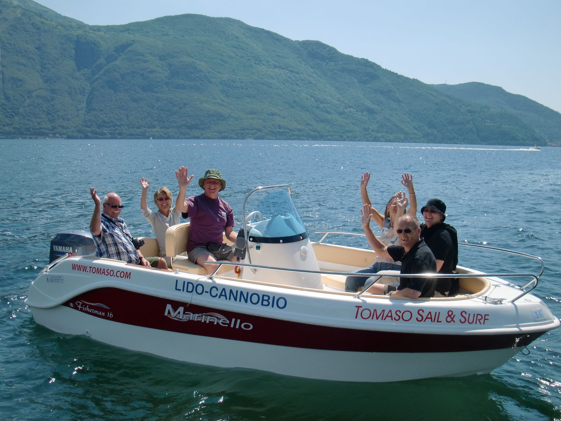 People waving from a small motorboat on a lake, with green mountains in the background.